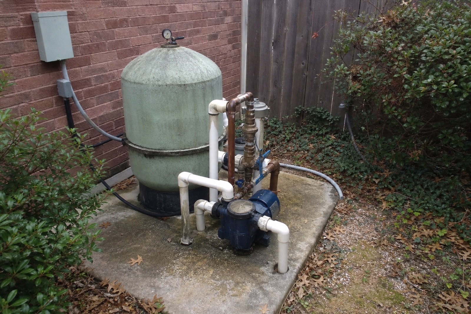 Residential pool equipment pad in a Plano backyard: aged filter tank, copper plumbing, navy pump.